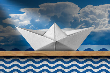 Closeup of a white paper boat on a wooden shelf at home, with blue waves painted on the wall and a sky with cumulus clouds.