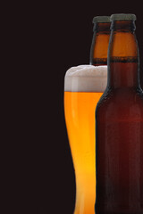 A frothy glass of pale ale between two brown bottles of beer, black background.