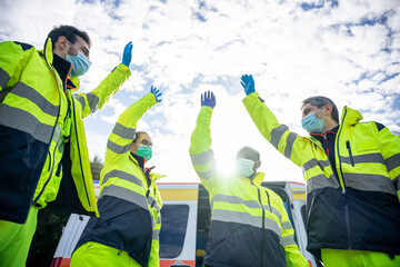 Portrait of a group of paramedics at the end of their shift in front of the ambulance while they high-fives in the air dressed in uniform and wearing a mask for protection from Coronavirus, Covid-19
