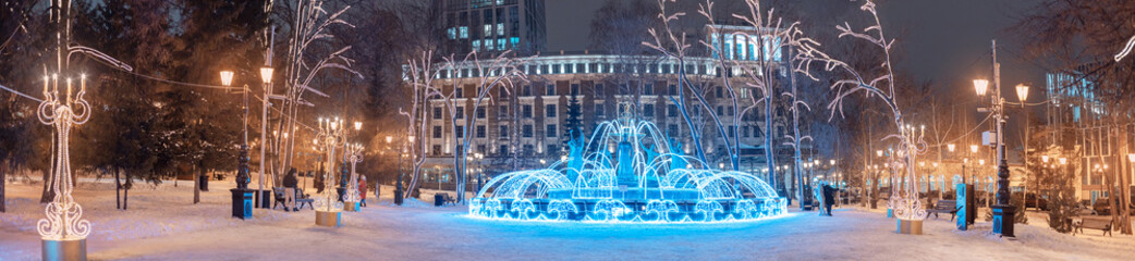 Wide panoramic view of the illuminated with garlands and twinkling lights fountain in the center of...