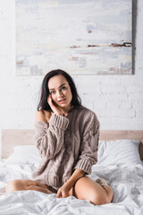  brunette woman in sweater and socks sitting in bed at morning