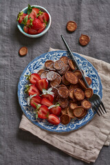 Mini chocolate pancake cereal with strawberries for breakfast on gray old textile tablecloth. Trendy home breakfast with tiny pancakes. Top view.