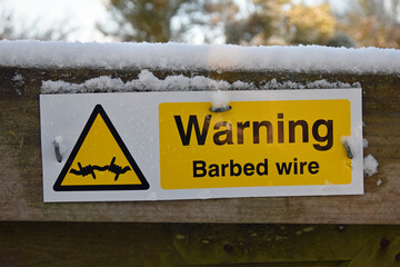 Yellow, black and white rectangular barbed wire warning sign with barbed wire icon in triangle. On...