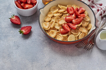 Mini white pancake cereal with strawberries in frying pan for breakfast on grey background . Trendy home breakfast with tiny pancakes. Top view.