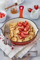 Mini white pancake cereal with strawberries in frying pan for breakfast on grey background . Trendy home breakfast with tiny pancakes. Top view.