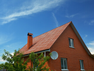 Fototapeta na wymiar A house of simple construction built from red bricks with a metal gabled roof, a chimney, roof gutters, an attic window, and a satellite dish antenna against the blue sky.