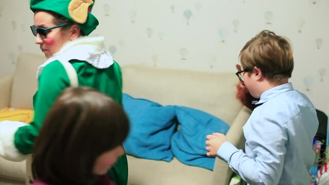 A teacher in an elf costume puts glasses over his eyes. The boy passes the object with his hands. The actor takes the children out of the room.