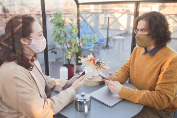 Two friends in protective masks discussing online apps on their mobile phones while sitting in coffee shop