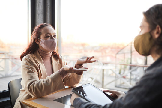 Two Young Business People In Protective Masks Discussing Future Plans During Business Meeting In Cafe