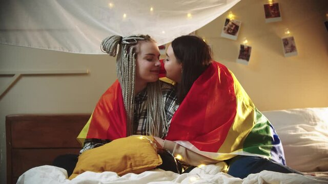 Young lesbian women sitting on the bed covered with LGBT flag - rub their noses and being cute