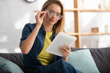  blonde woman touching eyeglasses while looking at digital tablet at home on blurred background