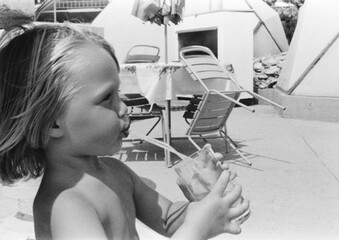 1976 vintage, seventies, retro monochrome image of a young girl with blond hair holding a glass with both hands drinking lemonade through a straw on a sandy sunny beach.