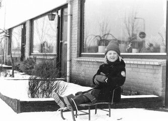 1978 vintage, seventies, retro monochrome portrait of a young girl with blond hair and duffle coat on a sledge in a winter snow setting in a suburban area.