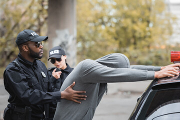 african american policeman frisking hooded offender leaning on patrol car near colleague on blurred...