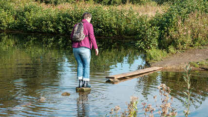 Frau in Gummistiefel überquert einen Fluss über einen kleinen Damm aus Steinen und Brettern - obrazy, fototapety, plakaty