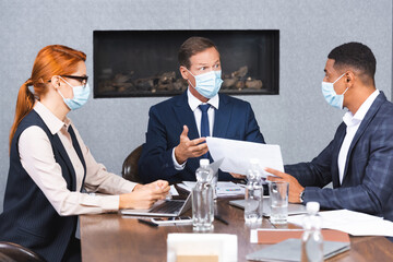 Multicultural businesspeople in medical masks talking while sitting at workplace with devices and papers on blurred foreground.
