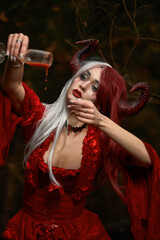 Maleficent Woman in Red Clothing and Horns in dark Forest. Posing with wineglass.