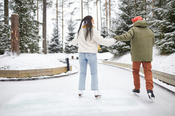 Fototapeta na wymiar Rear view of two friends in warm clothing holding hands skating together in the park in winter day