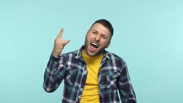 Happy adult guy having fun, enjoying music or event and showing rock n roll horns gesture, shouting yeah with joy, standing over blue background