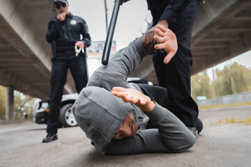 african american policeman with truncheon arresting hooded offender covering face while lying on street on blurred background.