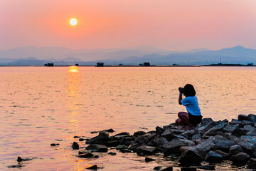 Young woman at waterfront take photos by DSLR camera beautiful nature landscape, sun on the bright orange sky over the mountains and lake water at sunset background, Krasiao Dam, Suphan Buri, Thailand