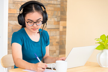 Asian teenage girl student with a headset is taking note learned online from school in a notebook using a laptop computer. Distance education from the class of the university by a video call from home