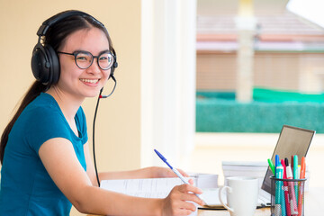 Young Asian teenage girl student with headset glasses is looking while  using a laptop computer study online from school distance education learning from a university class at home for background