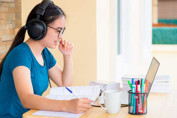 Asian teenage girl student with a headset and eyeglasses looks at laptop computer online learning from school and speak into a microphone. Distance education class of the college video call at a home