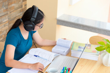 Young Asian teenage girl student with a headset glasses sitting on the desk reading a book using laptop computer online study from school. Education from the class of university by video call at home
