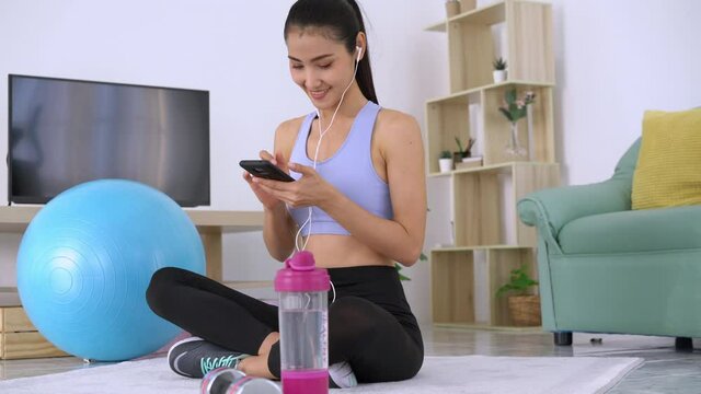 A beautiful young woman wearing headphones is using her smartphone while doing exercises sitting on the yoga mat in living room and listening to music on smartphone