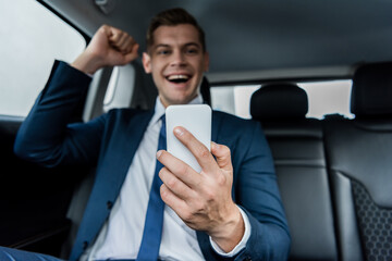 Smartphone in hand of cheerful businessman showing yeah gesture on blurred background in car.