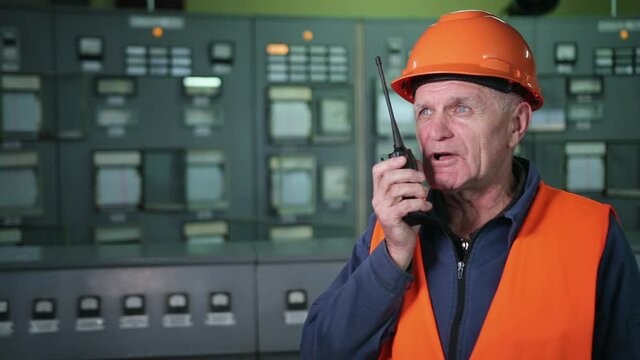 Adult male engineer in protective helmet, workwear using walkie talkie while standing in large workshop of contemporary factory. He holds vhf walkie-talkie radio hands. Industrial factory maintenance