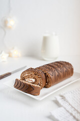 Brown chocolate sponge roll cake or roulade with white cream sliced on plate with knife, textile towel and glass of milk on white wooden table with garland lights at winter holiday. Vertical