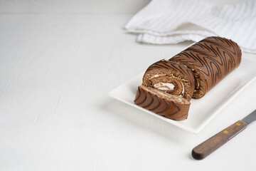 Chocolate sponge roll cake or roulade with white cream filling sliced on rectangular plate with kitchen knife on white wooden table with textile at winter holidays. Image with copy space, horizontal