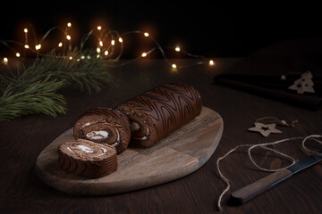 Chocolate sponge roll cake or roulade with white cream sliced on wooden cutting board with knife and warm garland lights on dark brown table at winter holidays with fir tree and towel. Horizontal