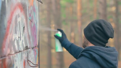 Youthful Guy Vandalizes the Wall Using a Spray Paint Can. Hooligan in Casual Clothing is Drawing Graffiti Outdoors. Youth Movement Crime Related with Street Art. Middle Close-Up View, Dynamic Shot