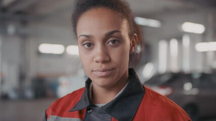 Tracking portrait shot of black female auto mechanic in uniform looking at camera while working in workshop