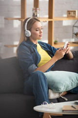 Obraz premium blonde woman in headphones texting on smartphone while sitting on couch on blurred background