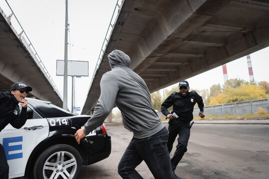 hooded offender running from multicultural police officers near patrol car on urban street.