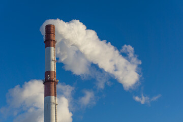A red and white chimney smokes against a blue sky. Smoking pipes of thermal power plant. Air emissions.