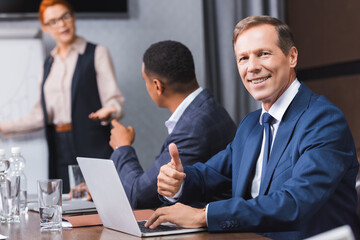 Smiling businessman with thumb up looking at camera near laptop with blurred multicultural colleagues on background.