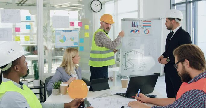 Likable confident professional multiracial team of engineers and managers gathering together in meeting room and brainstorming over joint building project