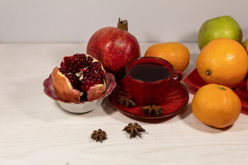 There is a red cup with pomegranate juice on a white table. Nearby are tangerines, apples, oranges, pomegranates and star anise.