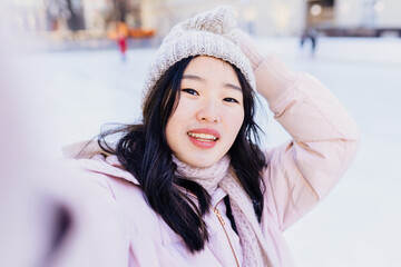 Outdoors lifestyle winter portrait of pretty young asian woman in powder pink jacket and warm knitted hat making selfie by the phone walking on the city street.