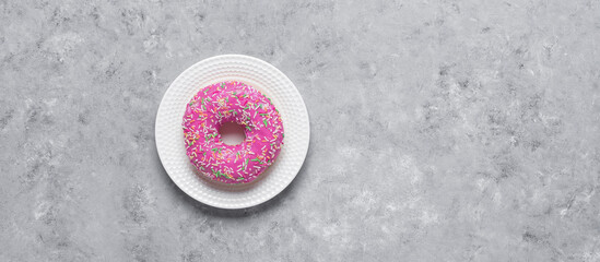 Obraz na płótnie Canvas Pink glazed donut on a plate, gray concrete grunge background. Donut with multicolored sprinkles, banner. Top view, flat lay.