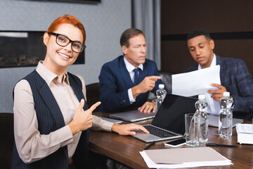 Smiling businesswoman looking at camera while pointing with finger at colleagues discussing on background.