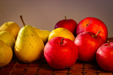 Several ripe yellow pears and red apples stand on a wicker basket illuminated by rays of the sun