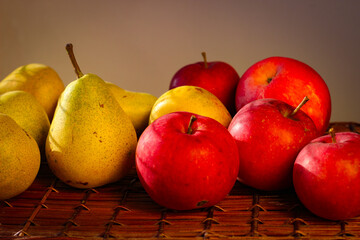 Several ripe yellow pears and red apples stand on a wicker basket illuminated by rays of the sun