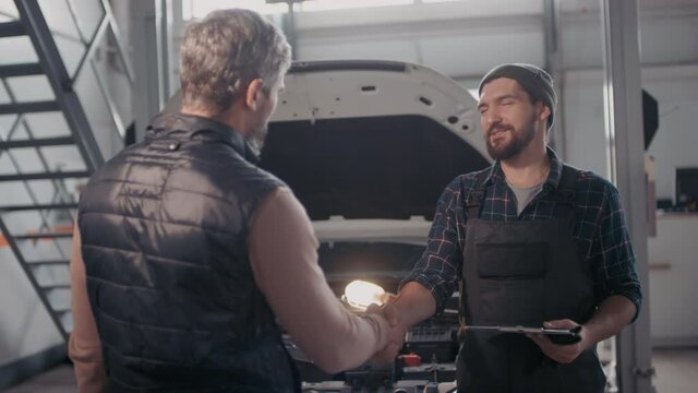 Slowmo shot of male client talking and shaking hands with cheerful auto mechanic with clipboard. Broken down car with open hood and portable work light inside is in background