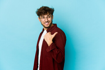 Young arab man on blue background points with thumb finger away, laughing and carefree.